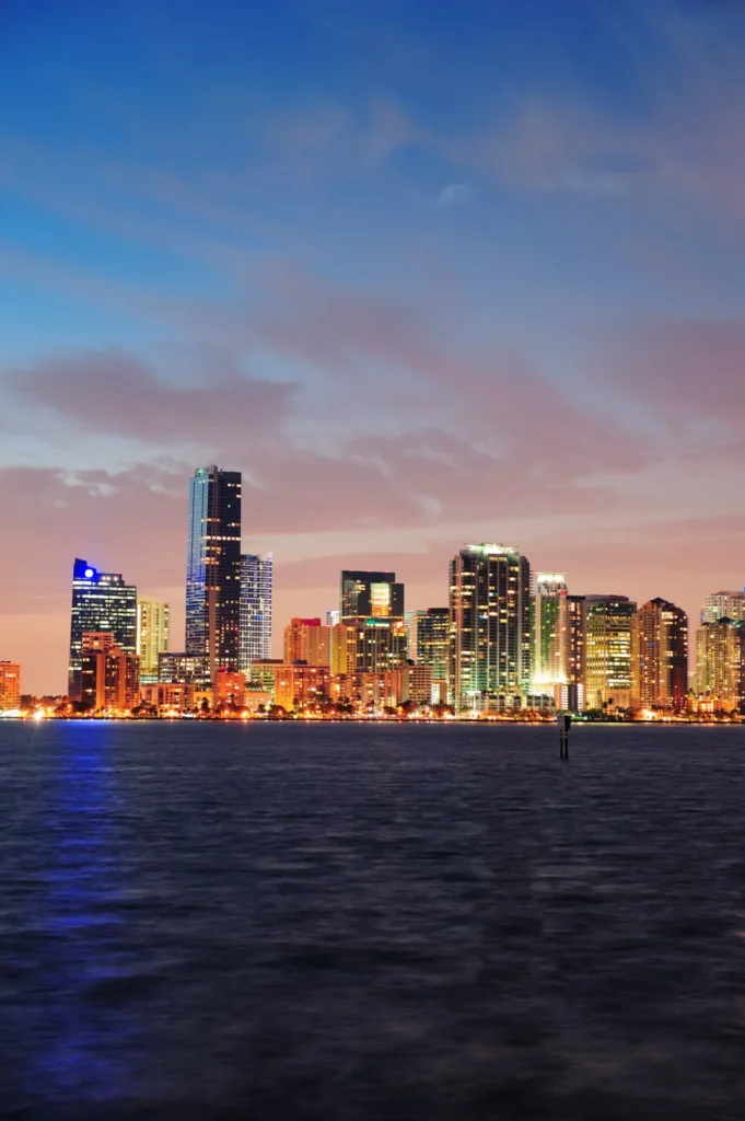 Skyline iluminado de Miami al atardecer sobre el agua, con rascacielos brillando bajo un cielo azul y rosado.
